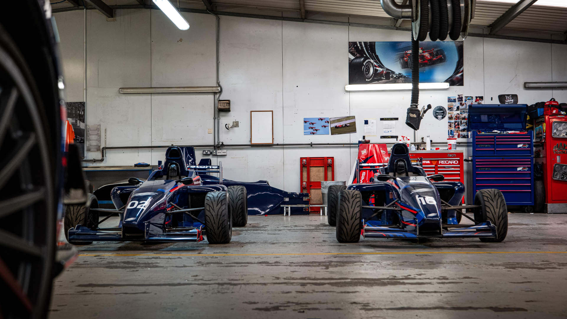 Two Formula Single seaters sit in the garage. Bodywork parts are visible in the background