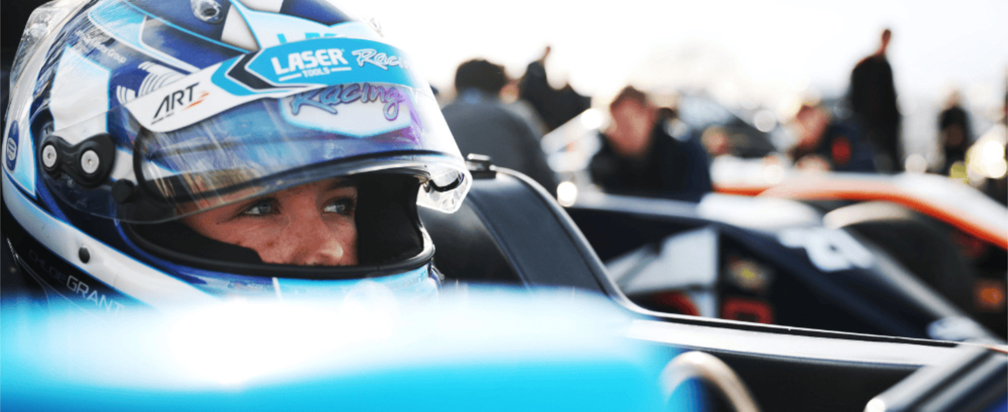Chloe Grant in her car, close up shot of wearing helmet with visor open, looking away at camera. Other cars can be seen in the background, suggesting this is the starting grid.