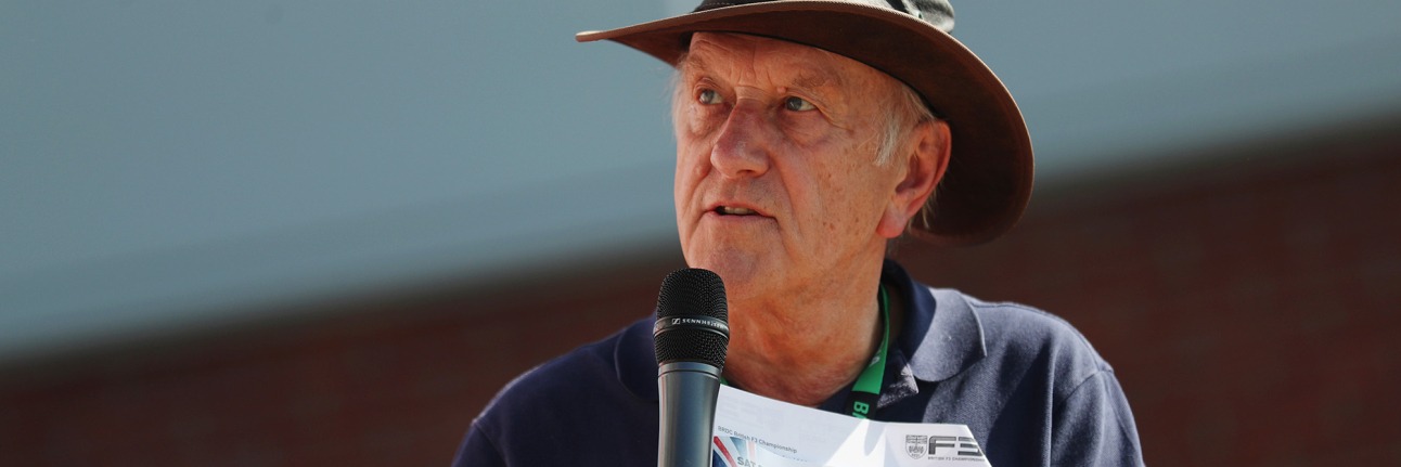 ian titchmarsh holds a microphone, looking off to the his left. Clutching a British GT programme and wearing a hat. 
