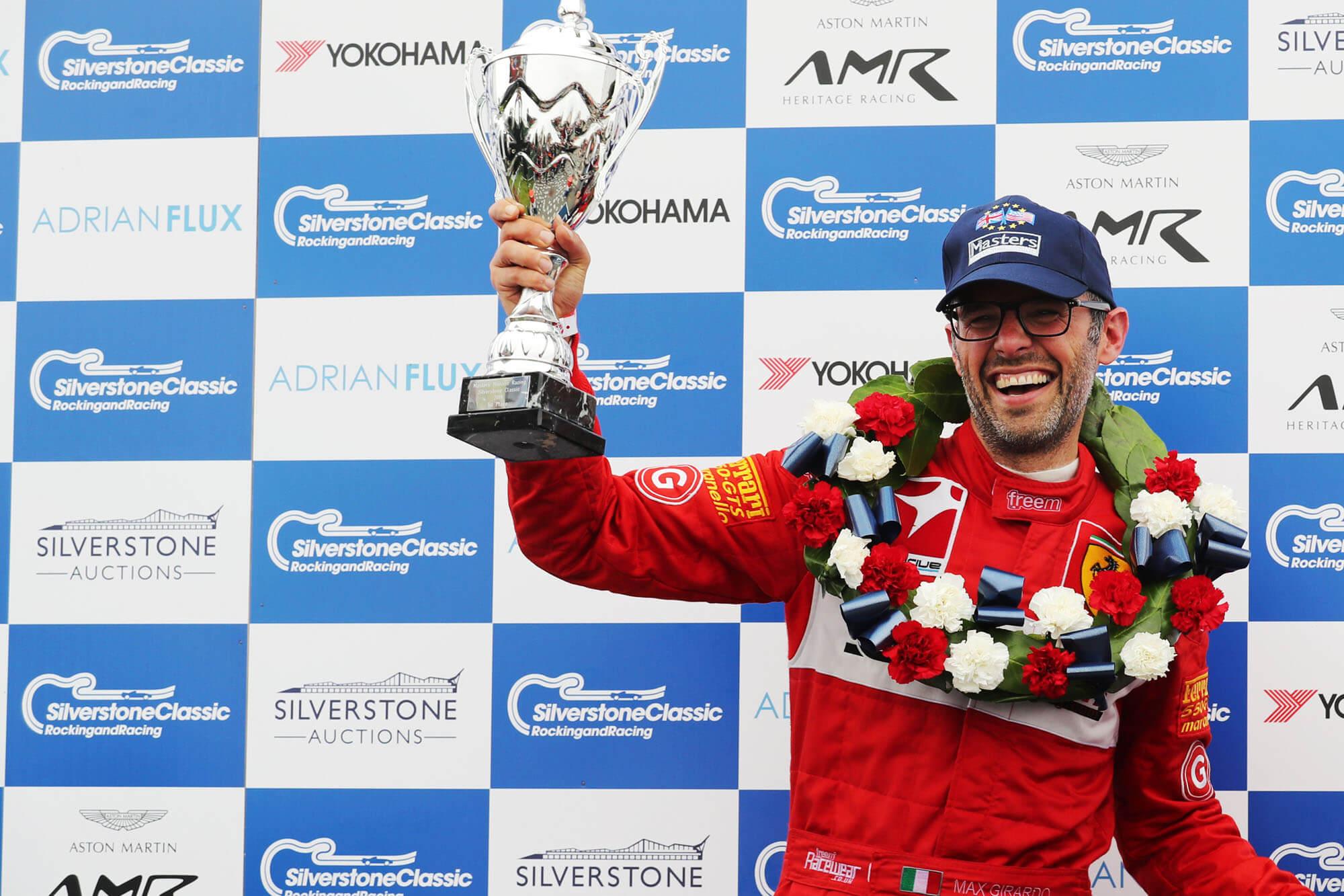 A race competitior standing on the winning podium smiling and holding up a winners trophy with a garland around his neck