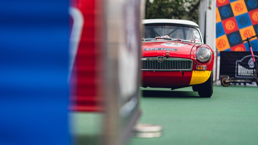 generations trophy car from motor racing legends sits half obscured behind a foreground object. MG car, red. 